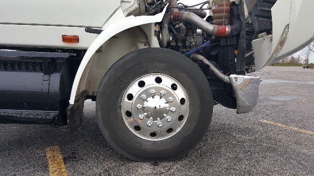 International 8600 front wheel and fender showing chrome hub with lug nuts, part of the front axle and suspension near open hood components.