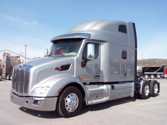 Peterbilt 579 truck in a light silver color, front three- quarter view, sleeper cab with high-roof; chrome grille with vertical bars and dual rear axles visible.