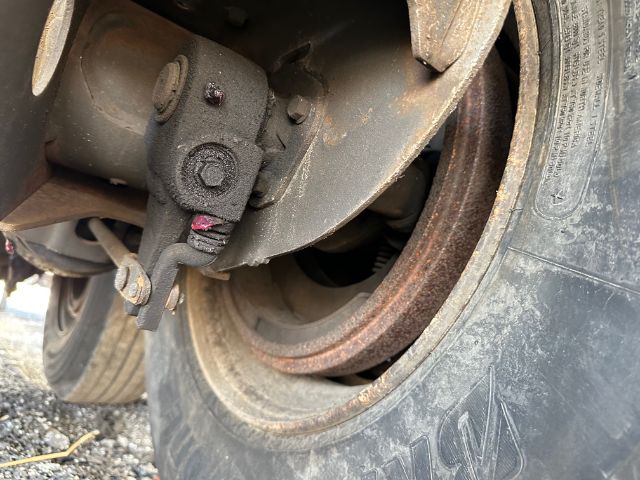 Trailer wheel and brake assembly with loaded drum, tire, and rusted hub. Rim shows circular opening and brake backing plate. Some dirt and wear visible.