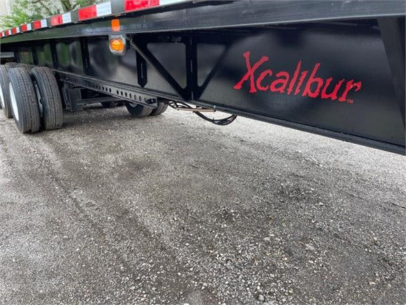 Flatbed trailer side showing Xcalibur logo in red on black frame, extendable 53'-90' undercarriage with dual rear axles and gravel surface.