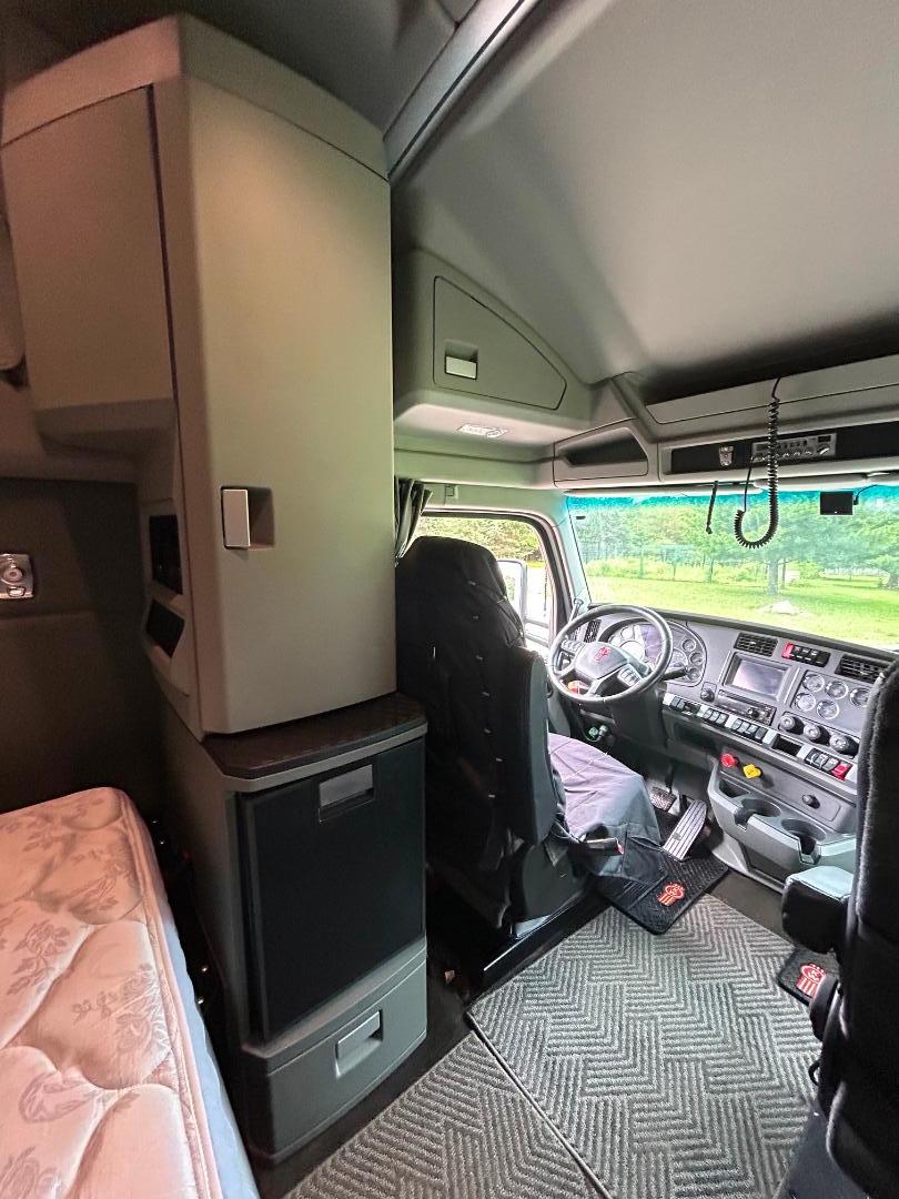 Kenworth T680 interior cab with driver seat, dashboard, and overhead storage; vertical cabinet and refrigerator area visible on left; patterned cab floor mat.