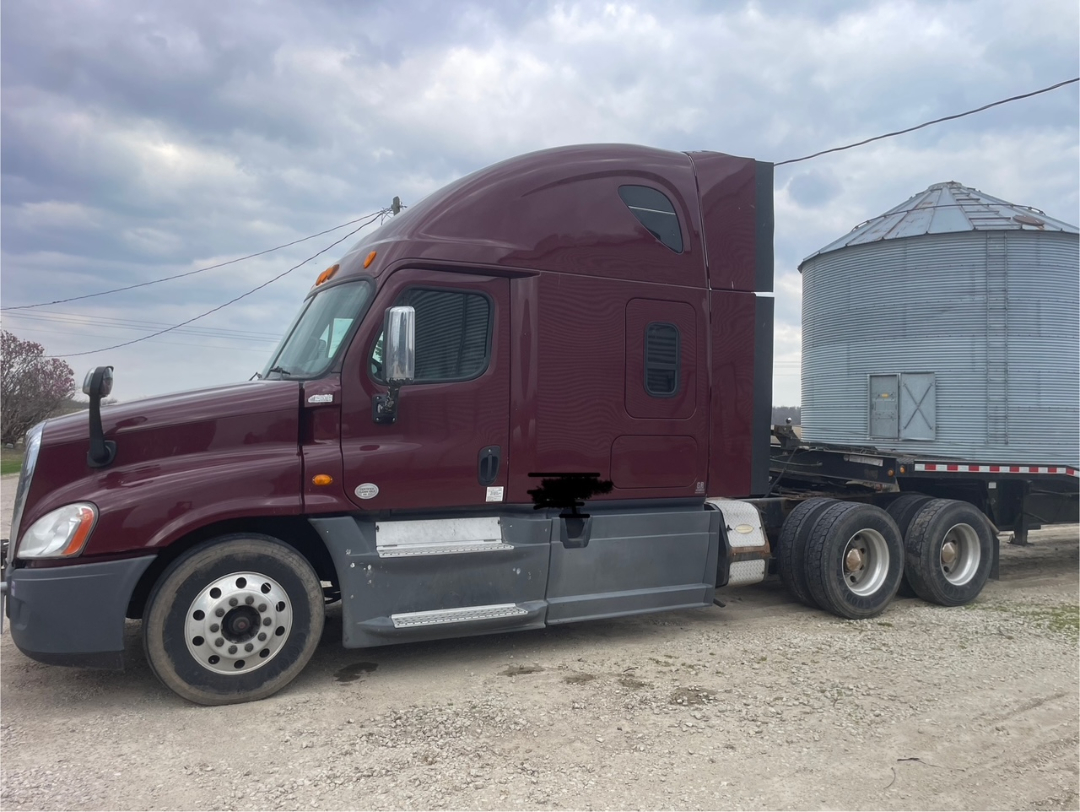 Maroon 2014 Freightliner Cascadia 125 conventional sleeper tractor in a gravel lot with a silver grain silage trailer attached.