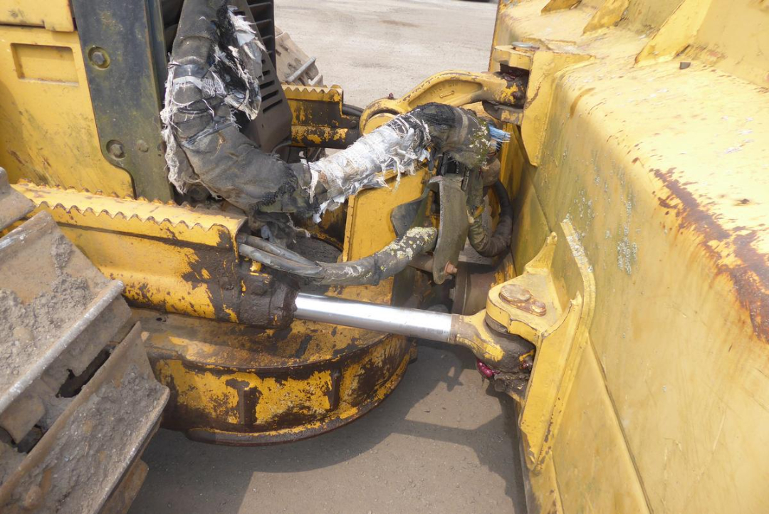 John Deere 650HLT dozer undercarriage and hydraulic cylinder with worn yellow paint, exposed hydraulic hose, and a shiny silver piston rod.