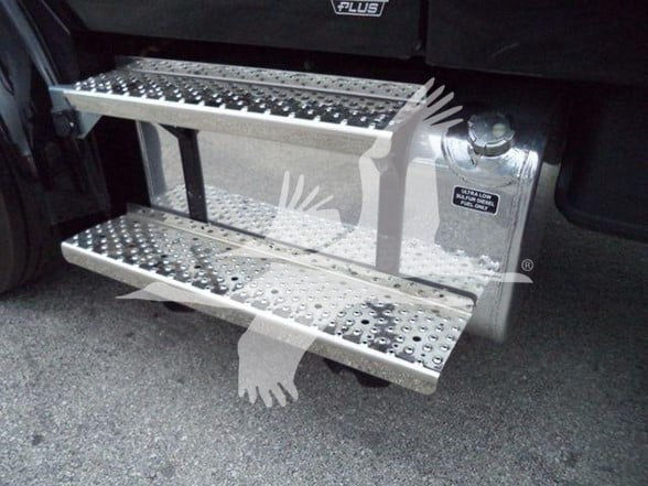 Platform steps attached to a Freightliner truck with perforated metal tread and a small label on the side near a hydraulic reservoir cap.