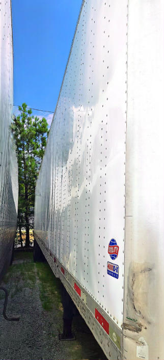 Trailer side showing perforated aluminum panels with small rivet holes; utility trailer with two logos/stickers near bottom; blue sky and trees visible in background.
