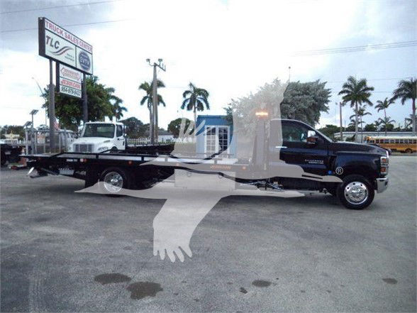 Tow truck with a flatbed, Chevrolet Silverado 6500HD image showing a black cab and bed, with palm trees and dealership sign in the background.
