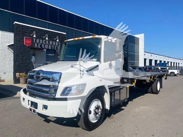White Hino flatbed truck in dealership lot, front-left view showing cab and metal flatbed with dual rear wheels.