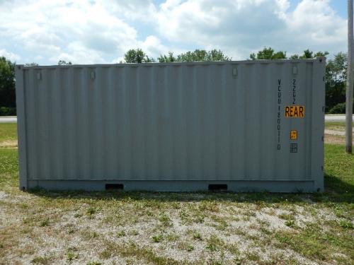 20' open side shipping container in a gravel lot. On the right end, vertical text includes 'REAR' in orange and other stenciled markings. Clear blue sky with some clouds.