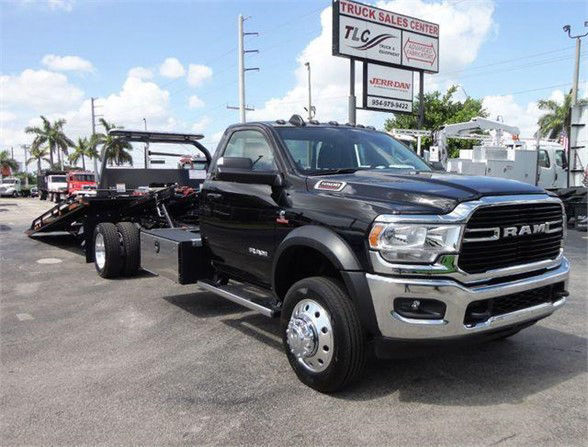 2024 Ram 5500 SLT tow truck with flatbed trailer attached, black cab, chrome grille, RAM emblem, dealership signs in background.
