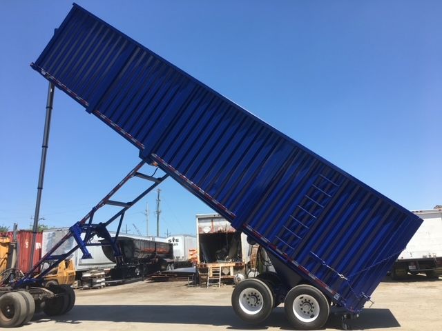 Utility dump trailer with blue box raised on hydraulic hoist and dual rear wheels, tilted for dumping.