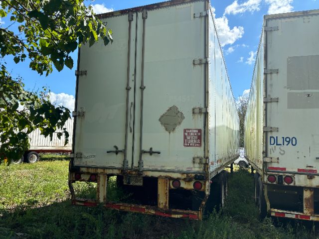 Rear view of a white Wabash trailer with double doors, rear bumper, and rusted hitch. Small damaged area on left door, warning sticker visible. Other trailer on the right.