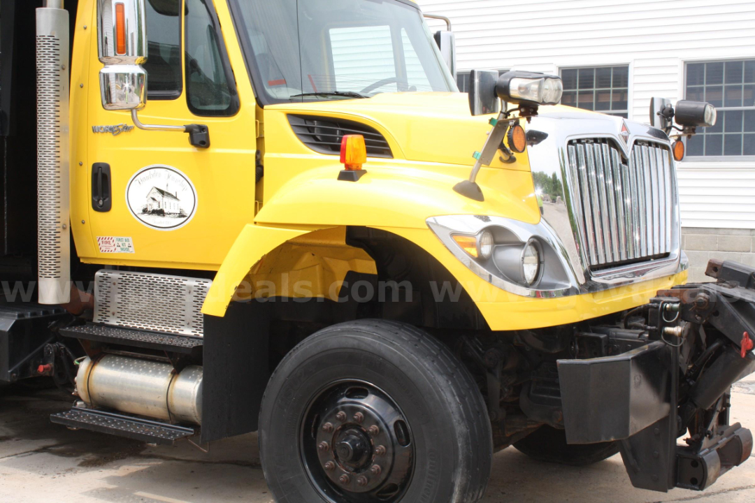 Front-left view of a yellow 2012 International 7400 plow truck showing front grille, headlight assembly, bumper, and large front wheel well.