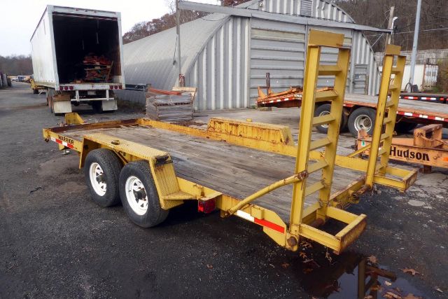 Trailer showing dual-axle flatbed with yellow paint and a ramps/vehicle-carrier area, parked on a wet lot with a pickup truck and a metal shed in the background.