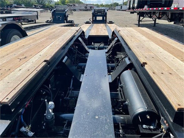 Lowboy trailer deck with wooden boards and center beam, hydraulic components, and trailer frame visible.