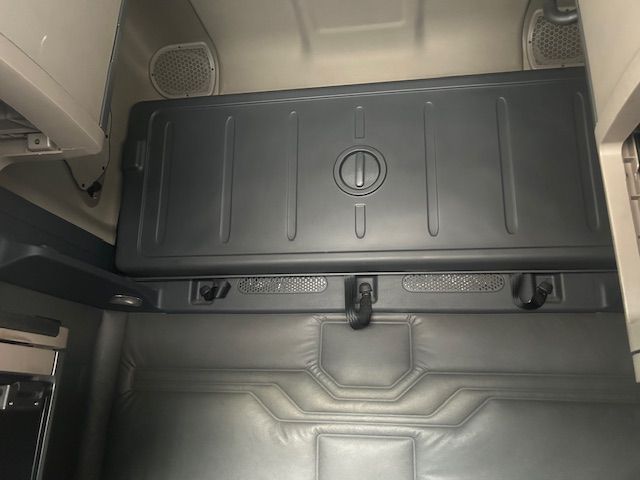Truck cab interior showing a storage box with a circular latch lid and a rubber floor mat with angular pattern. No visible labels or text other than the circular latch.