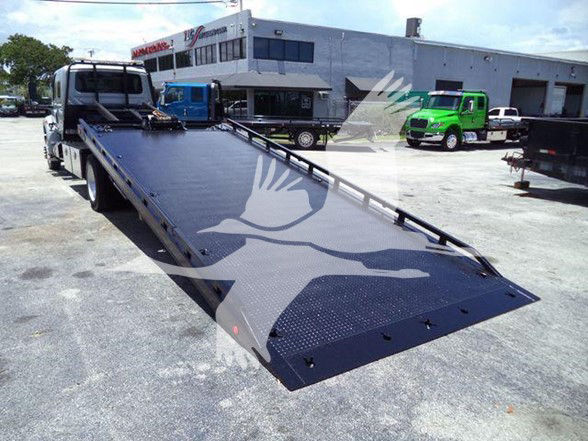 Flatbed tow truck platform with a long ramp extending toward the camera; warehouse and trucks in the background.