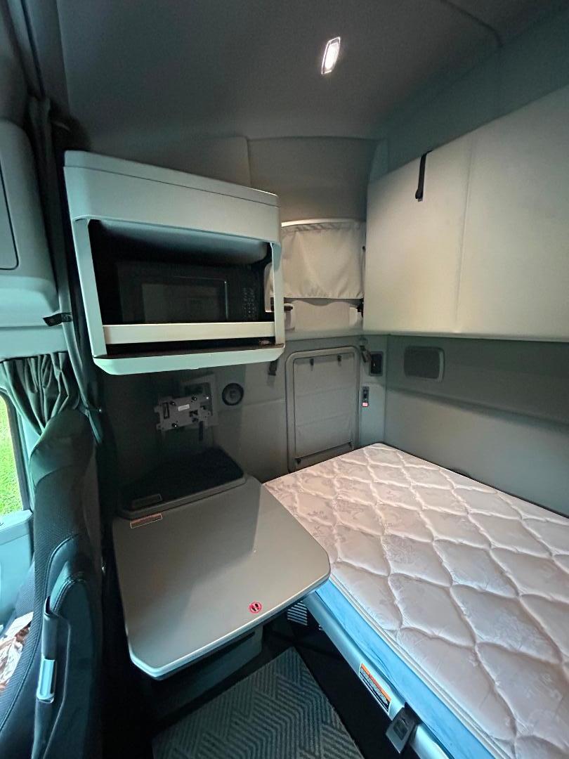 Truck sleeper cabin interior showing mounted microwave, foldout table, bed with mattress, and storage cabinets.