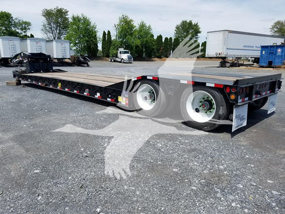 Trailer showing lowboy flatbed with dual rear axles, metal deck, and white wheels; branding and safety decals along side; background includes trucks and trailers.