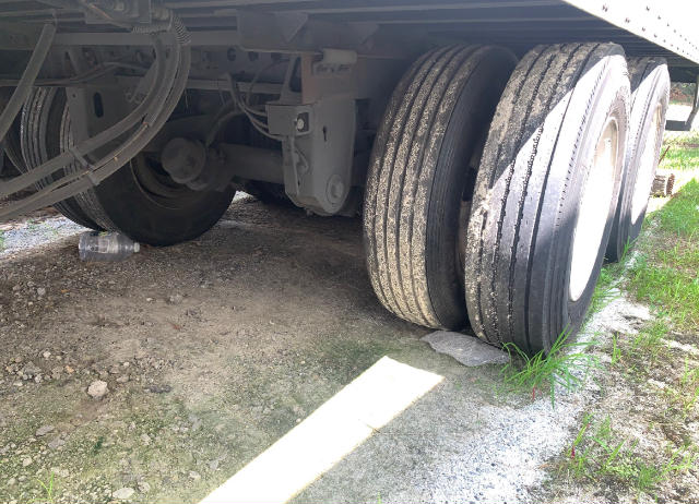 Trailer undercarriage with dual rear axles and tires, visible suspension components, frame rails, and gravel ground.