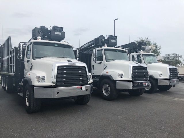 Three white Freightliner 114SD trucks lined up in a lot, grapple trucks with large front grilles and booms on top.