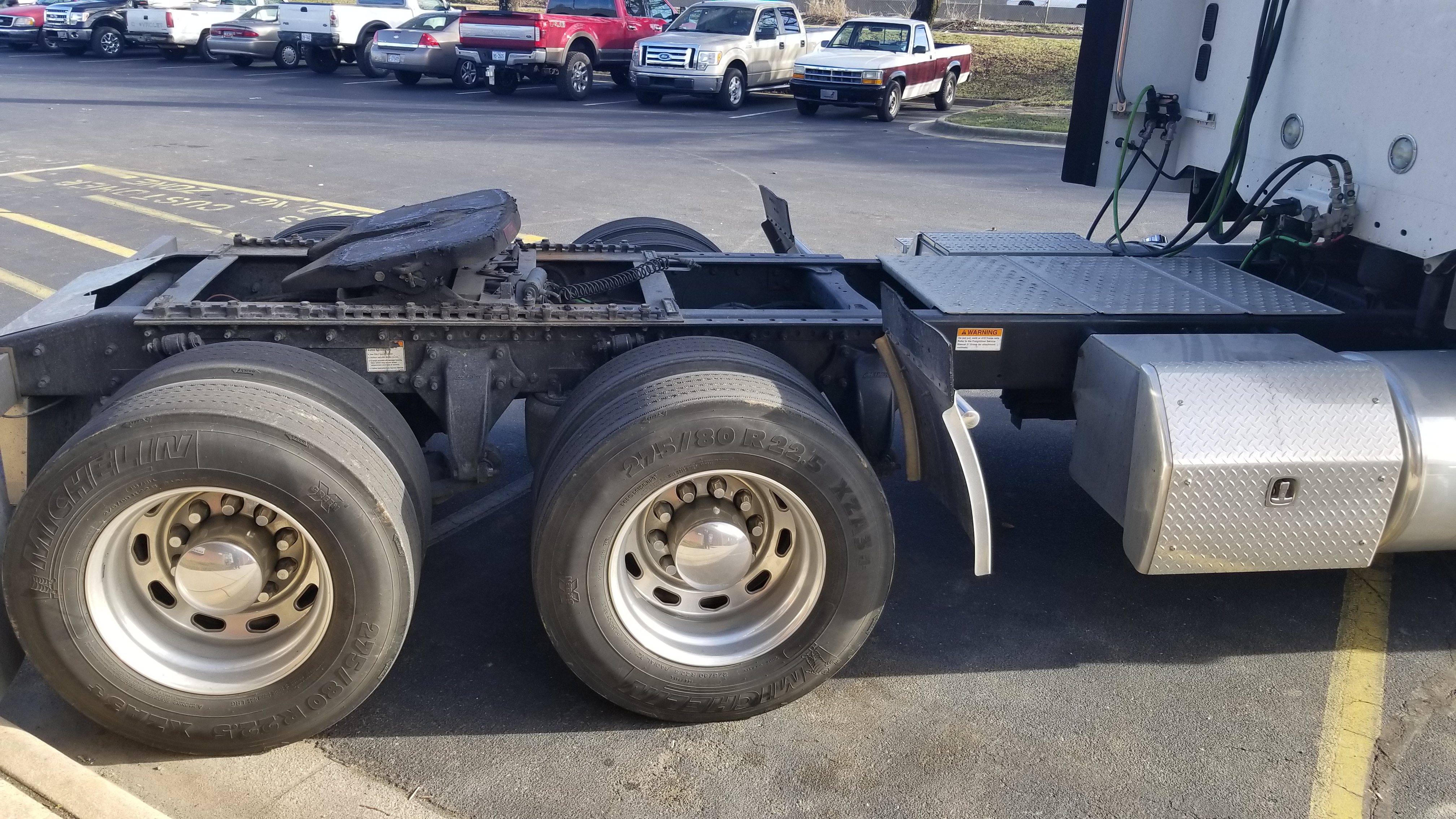 Tractor trailer chassis showing dual rear axles and Michelin tires, fuel/air tanks and diamond tread toolbox on the side, with cab connections in the background.