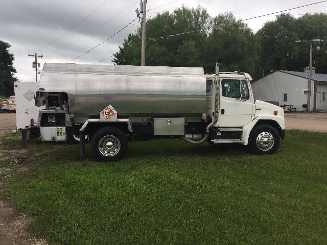 1999 Freightliner FL-70 gasoline truck with a large polished stainless steel fuel/tanker body. Visible are a white cab, chrome step, fuel tank, and hazard placard near rear wheel.
