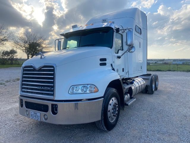Semi truck cab front-left side showing Mack grille, headlights, and bumper; large mirrors; side fuel tanks; two-axle day cab configuration; license plate visible on bumper.