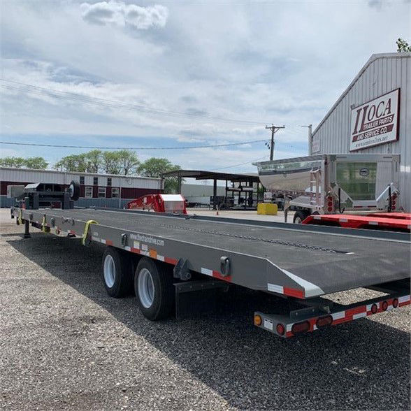 Trailer rear and side view of a 2024 drop deck container delivery trailer with tandem axles, gray flatbed, red/white reflective tape, in a lot near a building with JOCA sign.