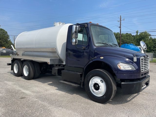 Truck with large cylindrical tank mounted on chassis. Front shows Freightliner-style cab, dark blue color, with round side mirror and headlights. The tank features access manholes and a metal diamond-plate skid along its base.