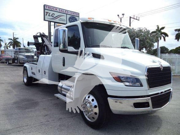 White 2025 International MV tow truck showing front grille and dual rear wheel chrome hubcaps; dealership lot background with palm trees.