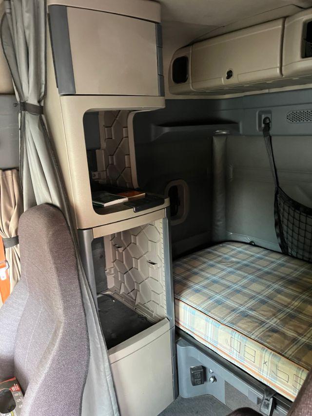 Truck sleeper cab interior showing bed with plaid mattress, overhead cabinets, and a small refrigerator/storage cabinet near the driver seat.