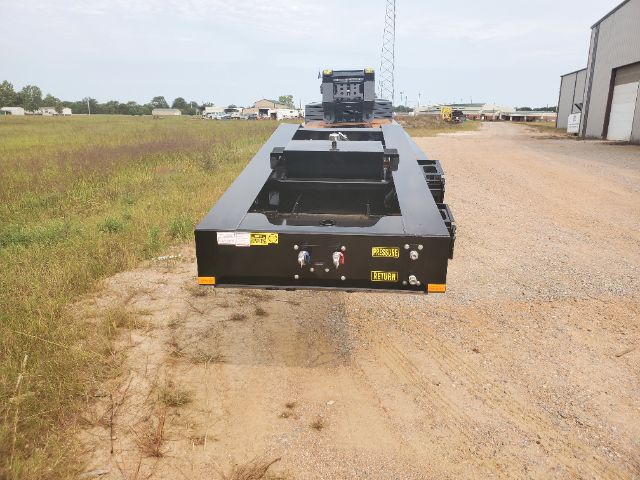 Flatbed lowboy trailer rear view showing control levers and safety decals on black steel surface.