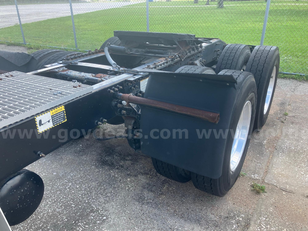 Chassis rear with dual rear axles and mud flaps. Visible components include a rusty bar, frame rails, and steel steps; warning label on frame; no engine, bare frame and wheels visible.