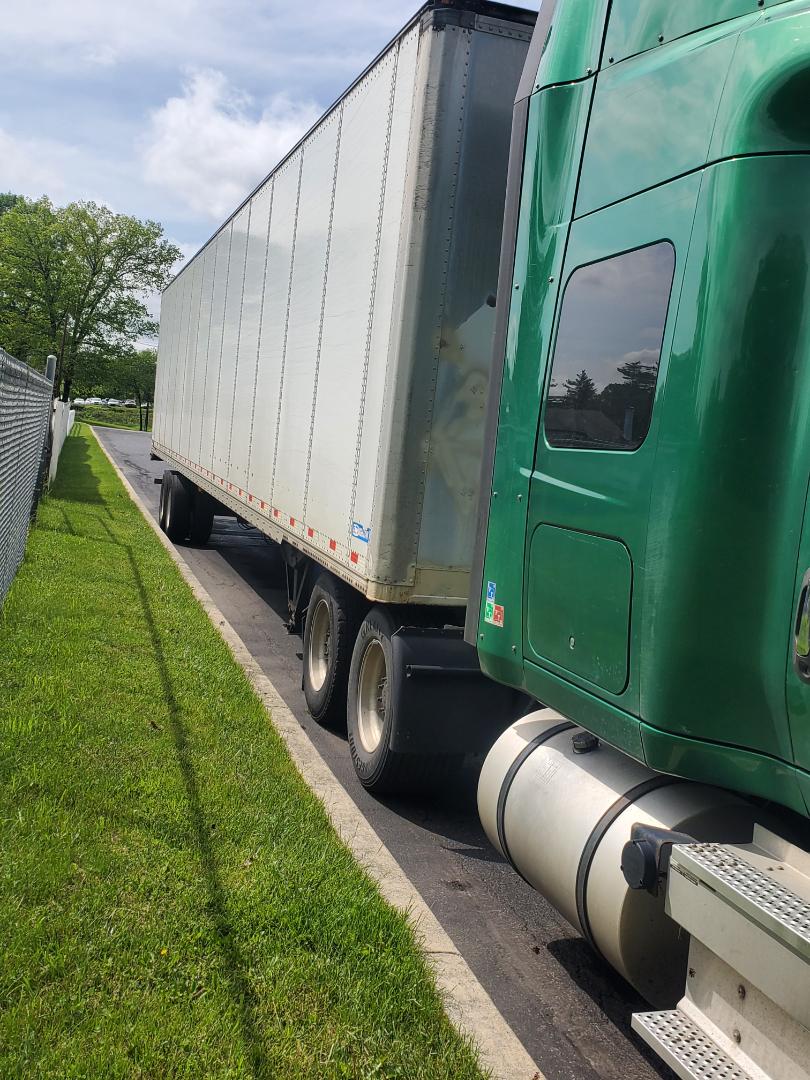Side view of a green 2019 International LT tractor connected to a long silver trailer, parked on a road shoulder with grass on the side.
