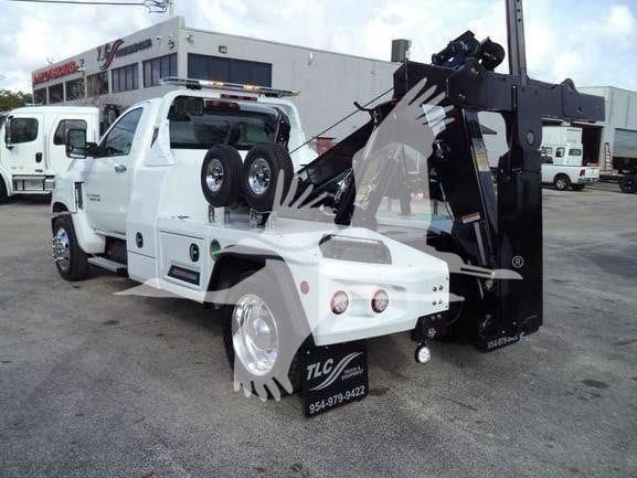 Tow truck with a front white cab and a black hydraulic crane/tower, spare tires mounted on the side, and TLC logo on visor; dealership in background.
