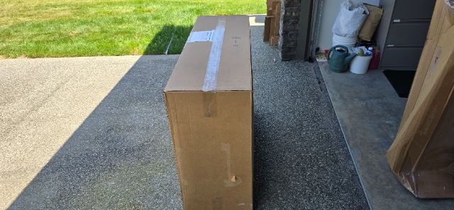 Box on driveway with tape seal, shipping packaging material visible, building doorway in background.