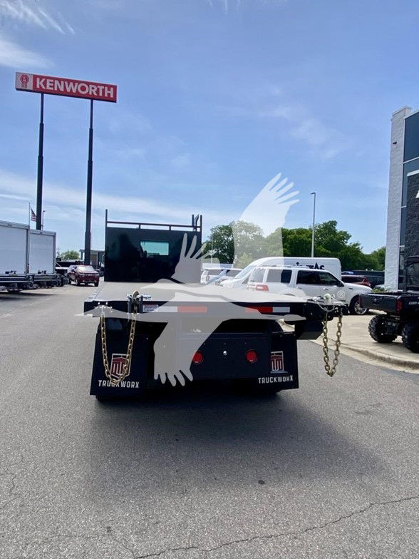 Kenworth dealership lot with a white flatbed Kenworth T480 seen from the rear; visible text includes KENWORTH sign, chains on the rear, tailboard, and mudflaps with logos.