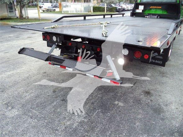 Flatbed tow truck rear with loading ramp extended, dual taillights, and hydraulic system visible.