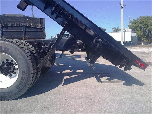 Truck body flatbed with a loading ramp lowered, behind the rear wheel shows a black flatbed trailer component and some red tail lights visible.