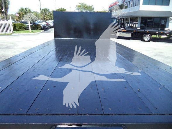 Flatbed truck bed with a large white silhouette of a bird logo, rear view shows flat wooden planks and a tall metal bulkhead, with a building and another flatbed in the background.