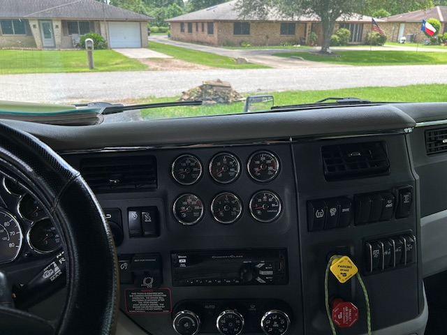 Panel with multiple gauges and controls from truck cab interior; left side shows switches and dials, center cluster has circular gauges, steering wheel on right, windshield reflected outside.