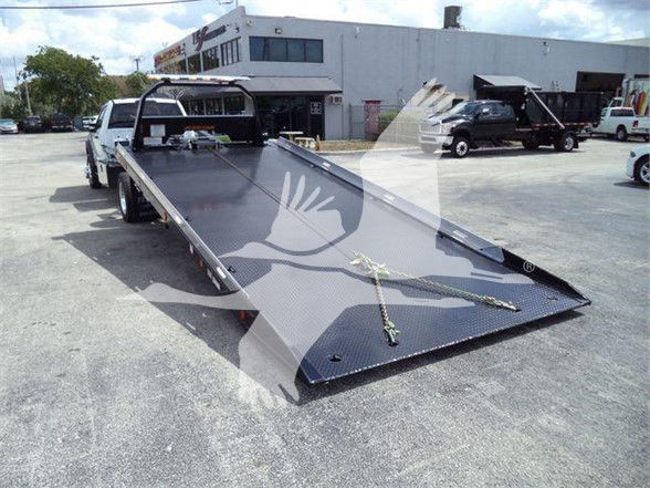 Flatbed tow truck bed on a new 2024 Ford F550, listed as stock J08894. Wide rear angle shows a black steel ramp with tie-downs; Ford towing equipment near dealership building.
