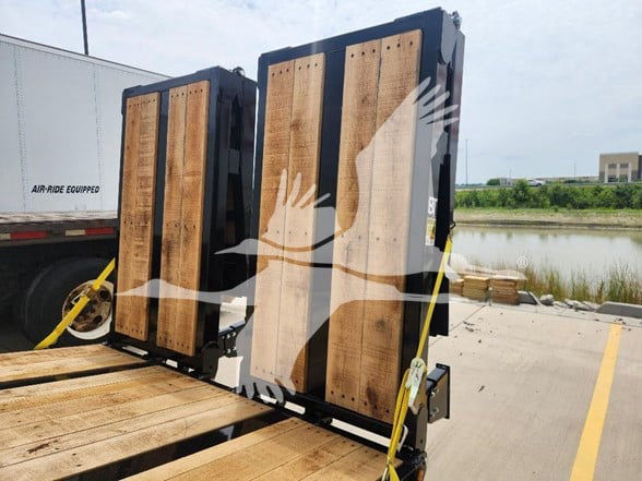 Flatbed trailer load with wooden decking and black metal frame, two raised ramps secured with yellow straps.