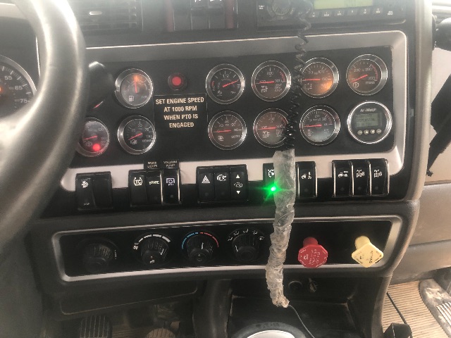 Dashboard of a Kenworth truck showing multiple gauges, switches, and warning labels. Text visible includes: 'SET ENGINE SPEED AT 1000 RPM WHEN PTO IS ENGAGED'. Various gauges with markings, and a green indicator light.