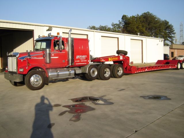 2007 Western Star 4900FA conventional sleeper truck with a red cab and a long red lowboy trailer, parked outside a building.