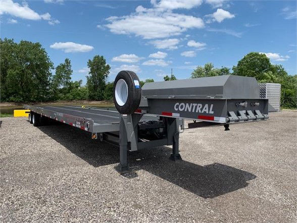 Flatbed trailer with tandem axle and a spare tire mounted on front, fade-gray deck and CON-TRAL equipment; clear blue sky and trees in background.