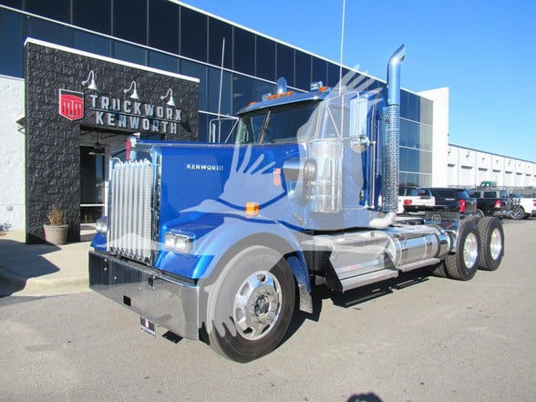Kenworth W900L conventional daytime cab truck in blue with chrome exhaust stacks and large grille, front view at dealership.