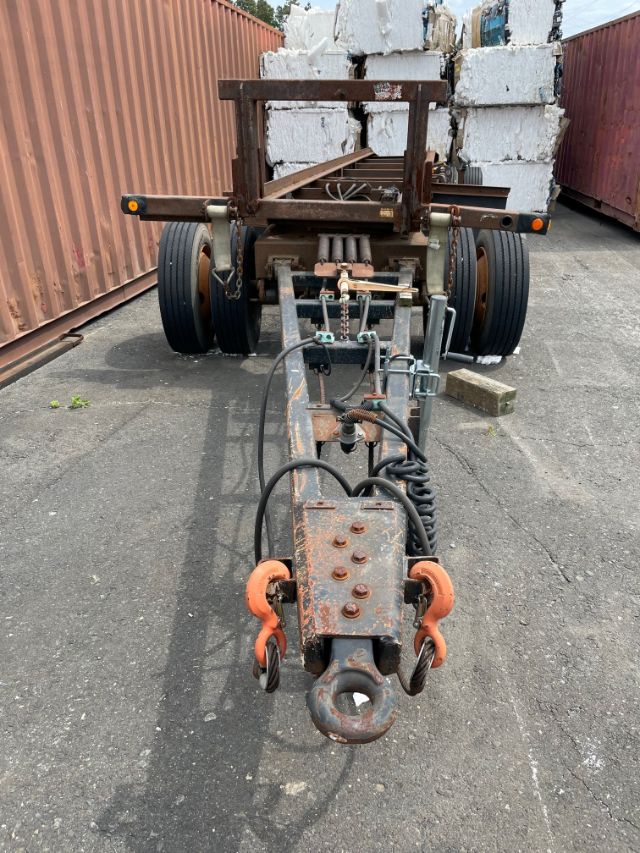 Trailer hitch assembly with hydraulic lines and coupler for a roll-off trailer, visible rust and support frame with orange hooks, rear dual wheels, and metal chassis.