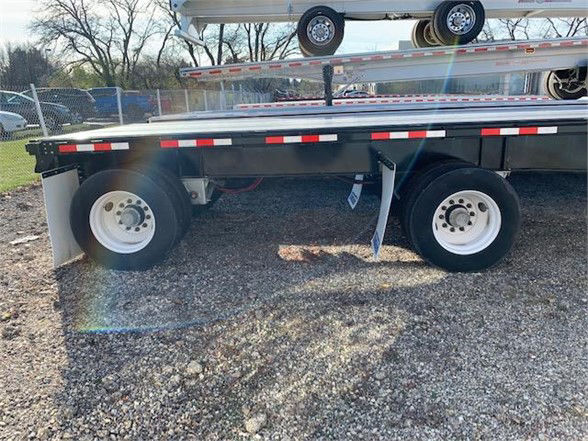 Flatbed trailer side view showing dual rear axles with white rims, steel deck, and reflective safety strips along edge.