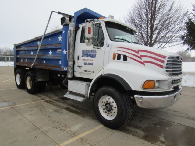 Dump truck with white cab and blue dump body featuring stars, side text including company name and numbers, front grille, and American flag graphics.